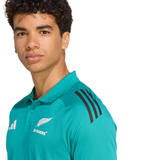 A person wearing the All Blacks 25/26 Training Polo by adidas, featuring teal fabric and black shoulder stripes, stands against a plain white background.