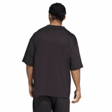 A person with braided hair is seen from behind, wearing a loose-fitting adidas All Blacks 25/26 Presentation T-Shirt and black shorts against a plain background.