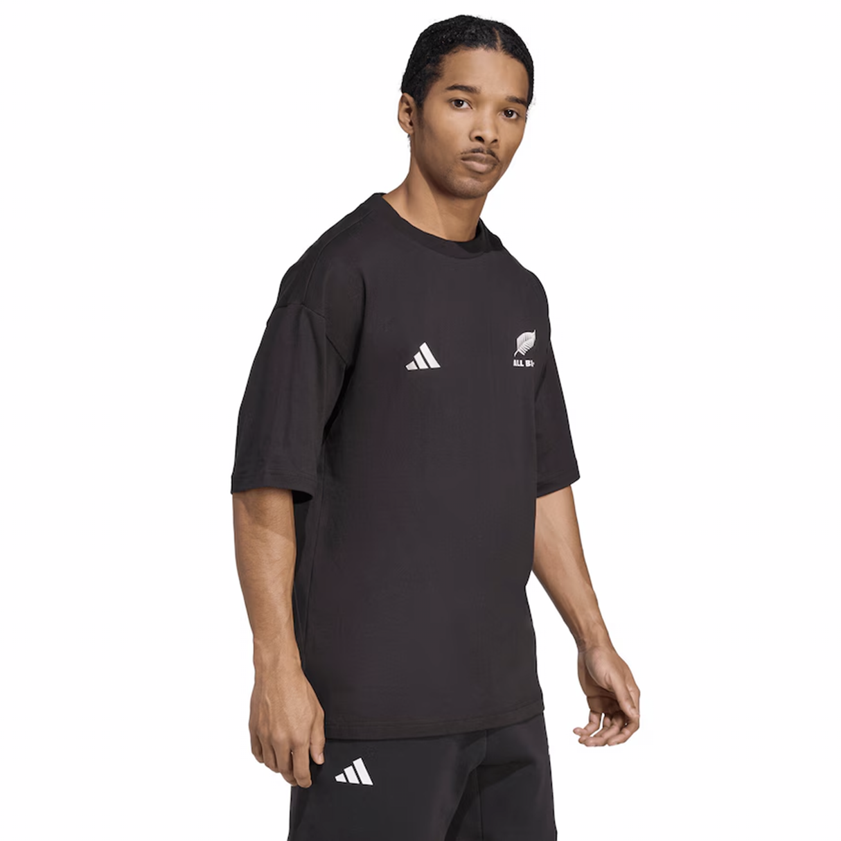 A man in an adidas All Blacks 25/26 Presentation T-Shirt, featuring white adidas logos and the iconic silver fern, stands against a plain background in shorts, looking at the camera.