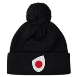 The Japan Shield Pom Pom Beanie by EMB Canterbury is black and features a unique logo of a red dot inside a white teardrop shape on the cuff, similar to the Japan Shield.