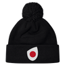 The Japan Shield Pom Pom Beanie by EMB Canterbury is black and features a unique logo of a red dot inside a white teardrop shape on the cuff, similar to the Japan Shield.