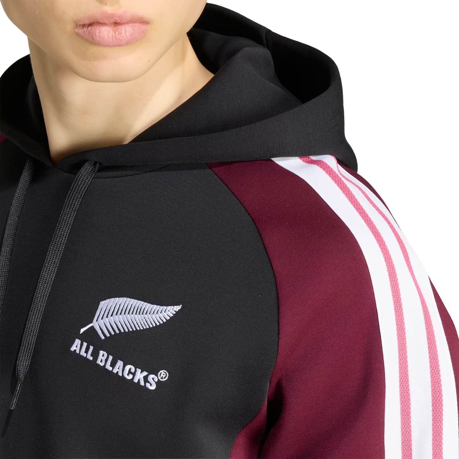 Wearing the adidas All Blacks 27/28 Hoodie in black and maroon, featuring iconic white sleeve stripes and a silver fern “ALL BLACKS” logo on the chest, this person proudly supports New Zealand rugby.