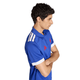 A person stands in profile, arms crossed, wearing the France FFR Home Supporters Replica Polo by adidas—featuring white shoulder stripes and red logos—displaying pride as a France FFR fan.