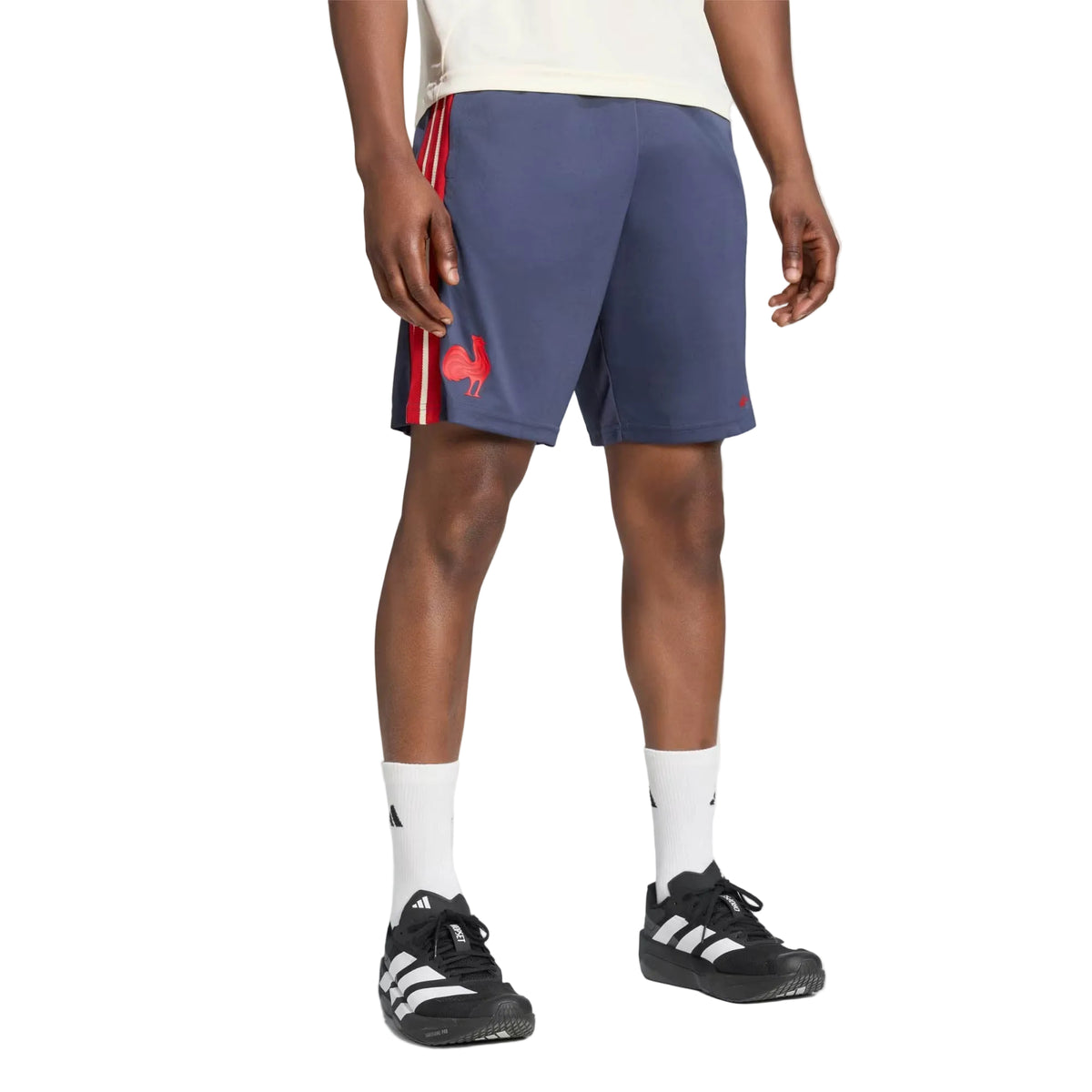 A person wearing the adidas France FFR 26/27 Gym Short, featuring a navy design with red stripes, paired with white socks and black-and-white sneakers, stands against a plain background.
