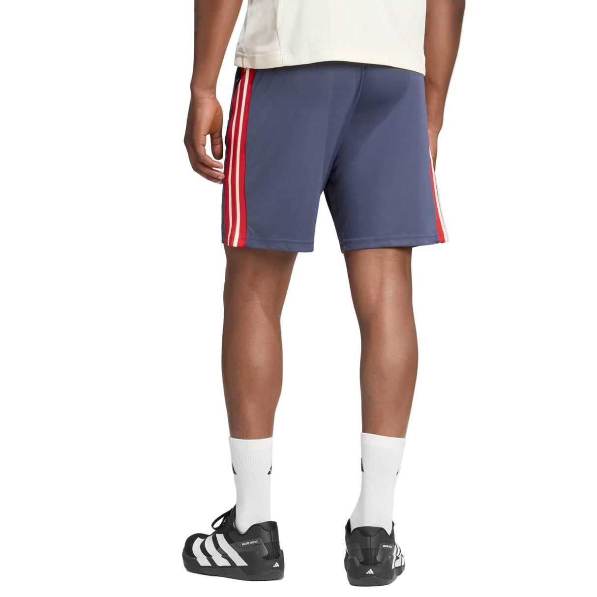 Person standing facing away, wearing adidas France FFR 26/27 Gym Shorts in navy with red and white side stripes, white socks, black adidas sneakers, and a cream T-shirt. Only the lower body is visible.