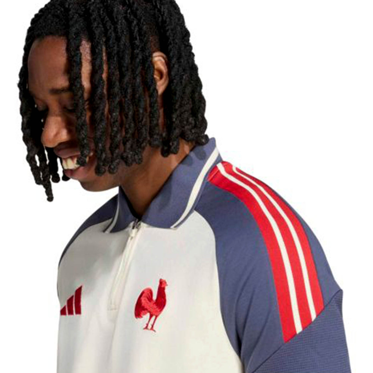A man with medium-length twisted hair smiles while wearing the adidas France FFR 26/27 Training Polo, featuring cream and navy colors with red stripes and a rooster emblem on the chest, set against a plain white background.