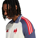 A man with medium-length twisted hair smiles while wearing the adidas France FFR 26/27 Training Polo, featuring cream and navy colors with red stripes and a rooster emblem on the chest, set against a plain white background.