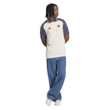 A person wearing the France FFR 26/27 Training Polo by adidas—a short-sleeve white and blue shirt with red accents—paired with blue jeans and white shoes, stands with arms behind their back against a white background.