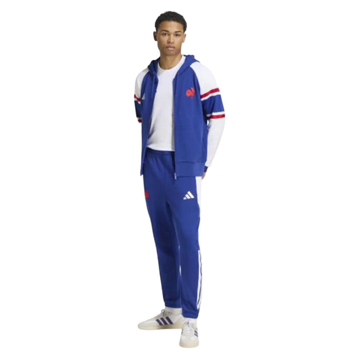 A person stands wearing a blue Adidas tracksuit with white and red accents, a white T-shirt, and white Adidas sneakers, layered with the France FFR 26/27 Travel Full Zip Hoodie by adidas for added comfort and style.