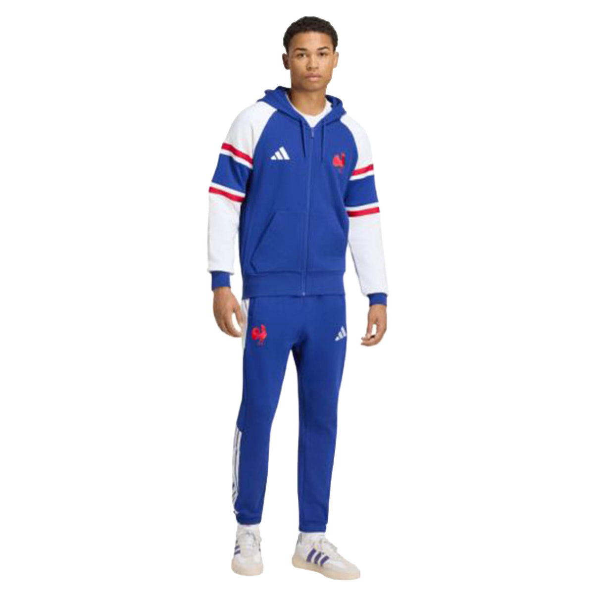 A person stands wearing the France FFR 26/27 Travel Full Zip Hoodie by adidas—a blue, white, and red tracksuit with a hood and matching pants. The look is completed with white sneakers and features adidas logo details on both the jacket and pants. This standout Full Zip Hoodie captures the dynamic style of adidas, making it perfect for France Rugby fans or anyone seeking comfort and sporty flair.