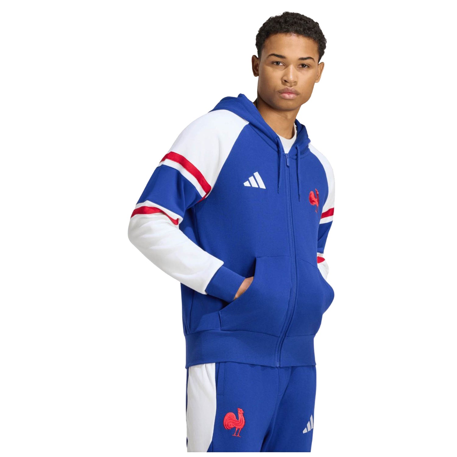 A man models the France FFR 26/27 Travel Full Zip Hoodie by adidas, featuring blue, white, and red colors with a rooster emblem and adidas logo, standing with one hand in his pocket against a plain white background and showcasing the vibrant style of this official France Rugby hoodie.