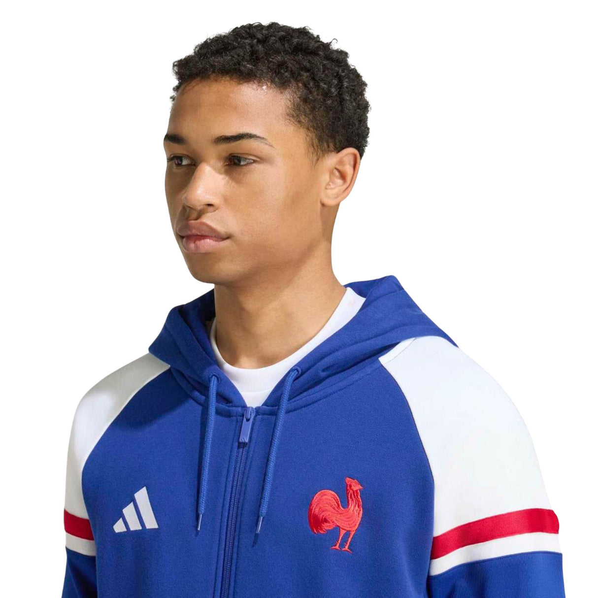 A young man wearing the adidas France FFR 26/27 Travel Full Zip Hoodie, featuring blue and white colors with red stripes and a rooster emblem, looks slightly to the side.