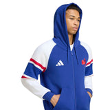 A person wearing the adidas France FFR 26/27 Travel Full Zip Hoodie, featuring a blue and white design with red accents, front pockets, and the adidas logo, stands with hands in pockets against a white background.