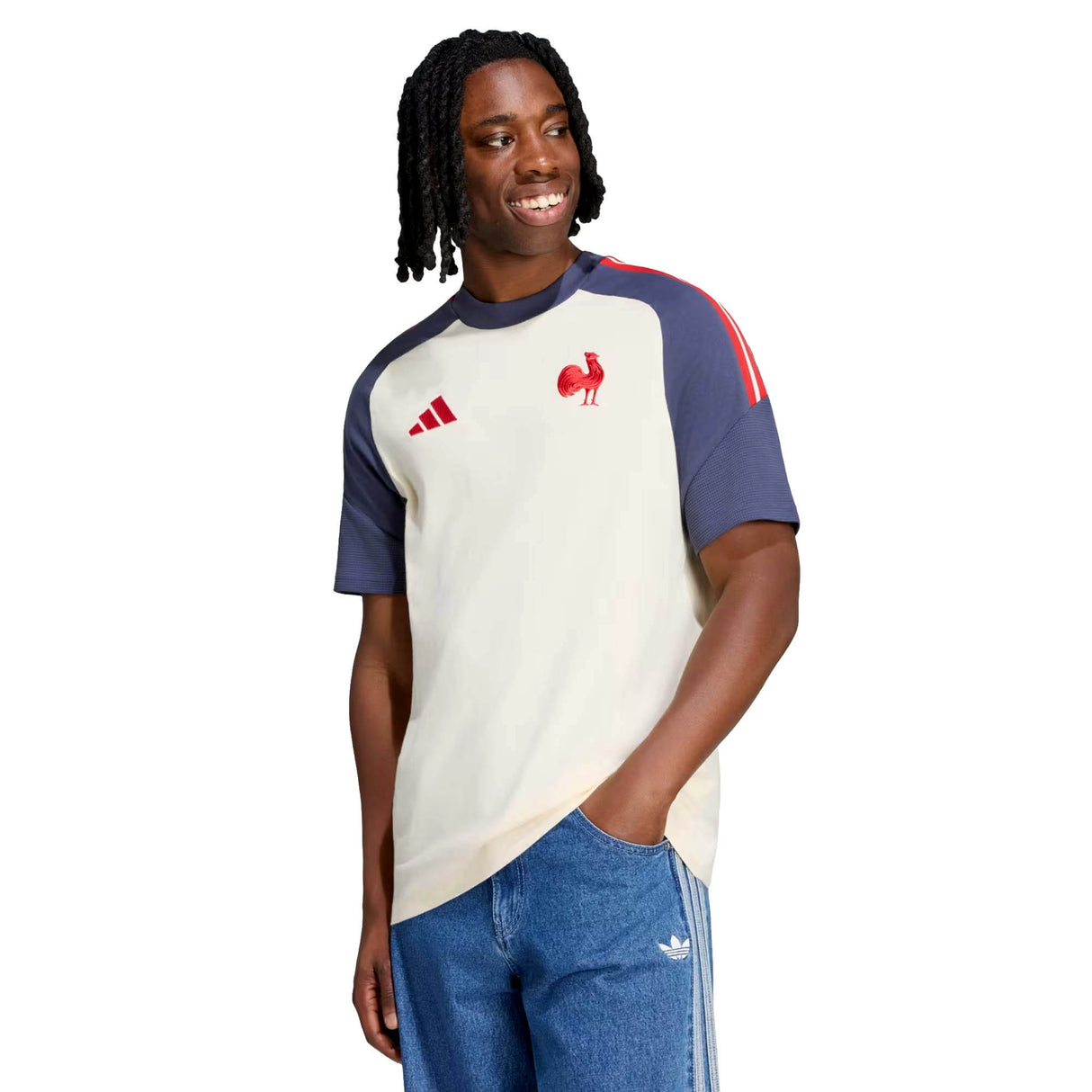 A person wearing the adidas France FFR 26/27 Cotton T-Shirt—featuring cream and navy colors with red accents—and blue jeans stands with one hand in their pocket, smiling and looking to the side.