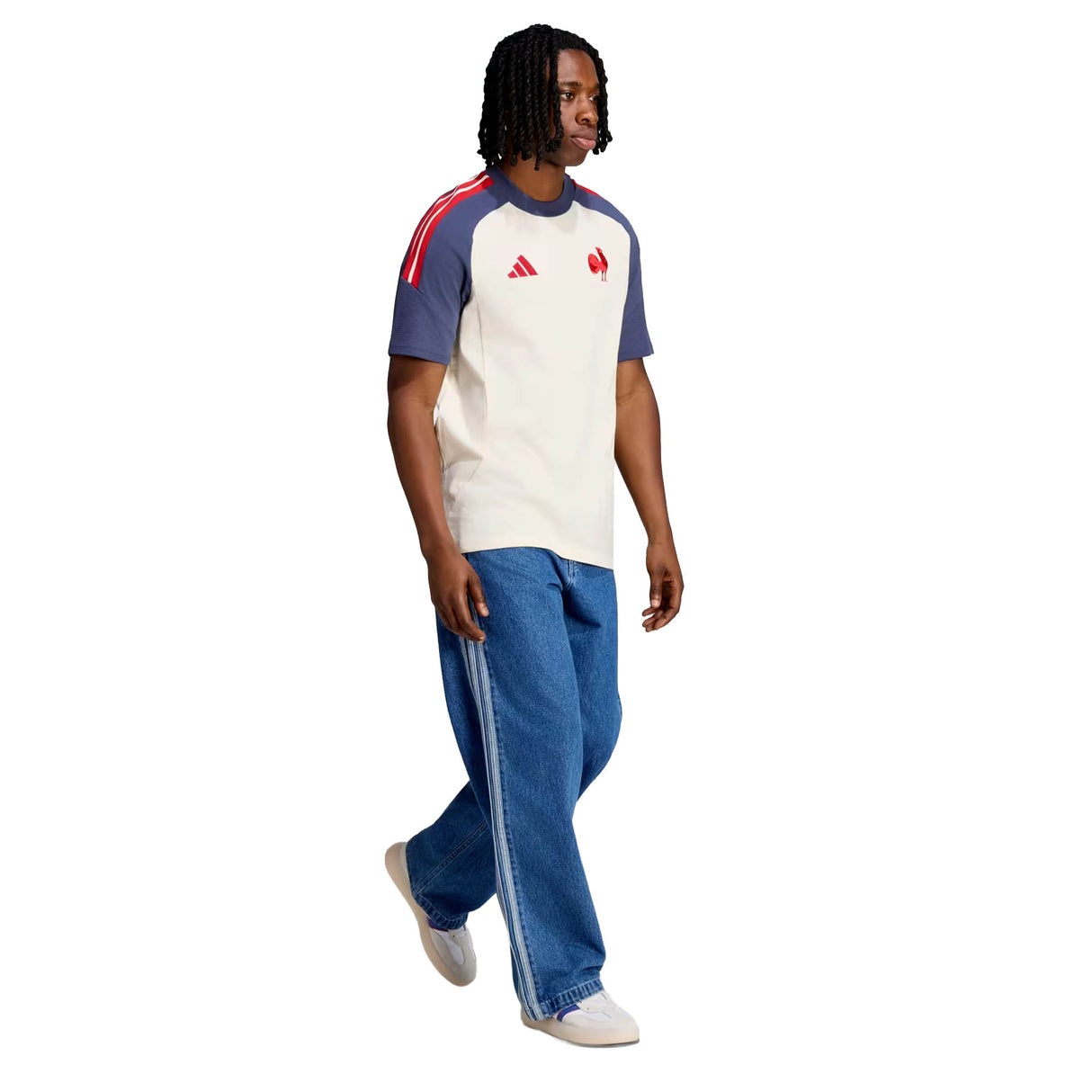 A person with medium-length braids wears the adidas France FFR 26/27 Cotton T-Shirt—white and blue with red accents and the Le Coq badge—paired with wide blue jeans and white sneakers, walking against a white background.
