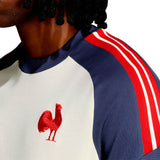 A person wearing the France FFR 26/27 Cotton T-Shirt by adidas, featuring a white and blue design with red shoulder stripes and the iconic Le Coq badge—a bold red rooster emblem—on the chest.