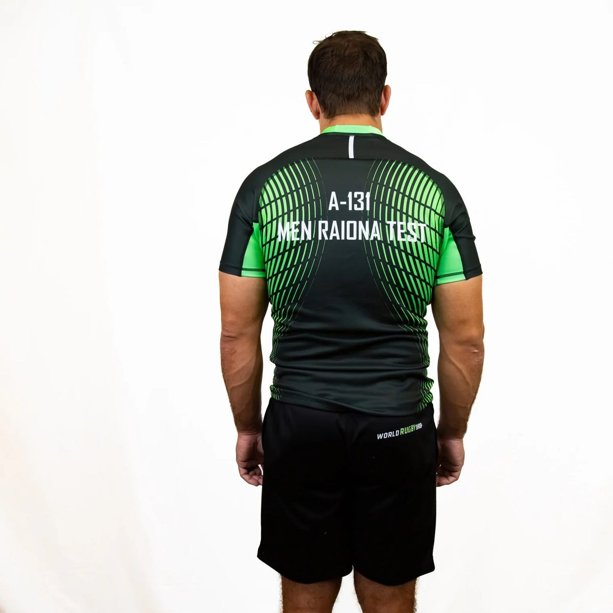 A man stands facing away from the camera, modeling the World Rugby Shop Test Product Jersey—a black and green athletic shirt with text—paired with black shorts, set against a plain white background.
