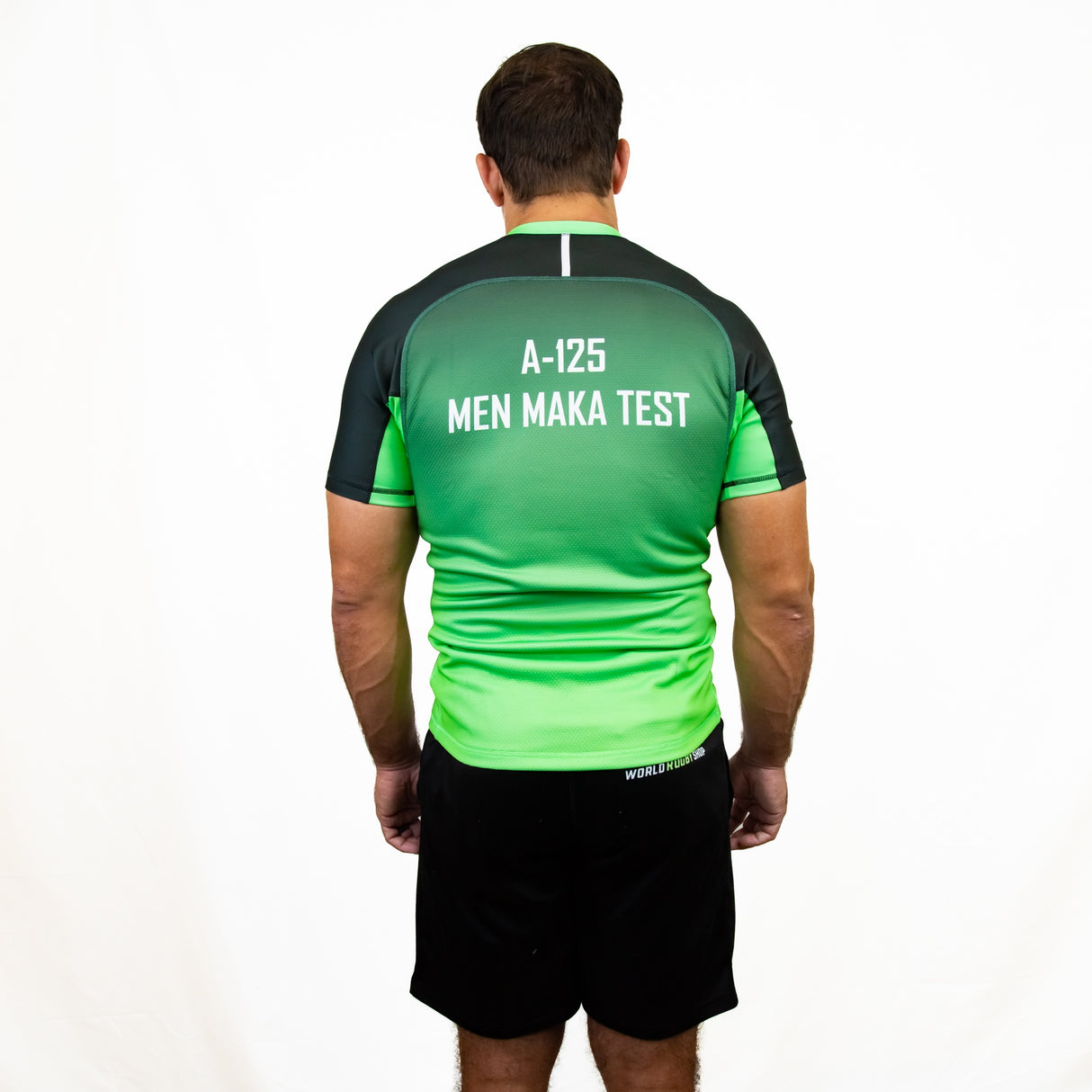 A man faces away from the camera, wearing a green and black Teamatical Canterbury Elite Full Custom Sublimated Jersey – Mens and black shorts, set against a plain white background.