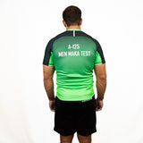 A man faces away from the camera, wearing a green and black Teamatical Canterbury Elite Full Custom Sublimated Jersey – Mens and black shorts, set against a plain white background.