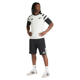 A person stands with hands on hips, wearing the adidas All Blacks 26/27 Travel T-Shirt, black shorts, white socks, and white sneakers against a plain background.