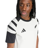A person wearing the All Blacks 26/27 Travel T-Shirt by adidas—a white and black short-sleeve shirt featuring shoulder stripes and two chest logos—facing slightly to the side.