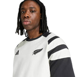 A person with long hair wears the All Blacks 26/27 Travel T-Shirt by adidas, characterized by its white and black color scheme and silver fern emblem, looking toward the camera.