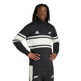 A person stands with hands on hips against a plain white background, wearing the adidas All Blacks 26/27 Leisurewear 1/4 Zip Sweatshirt in black and white.