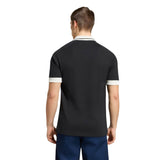 A man stands facing away, wearing the adidas All Blacks 26/27 Travel Polo with white trim on the collar and sleeves, paired with blue pants, against a plain white background.