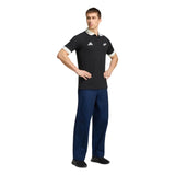 A man stands with hands on hips, wearing the adidas All Blacks 26/27 Travel Polo featuring team logos, along with blue pants and black shoes against a white background.