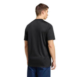 A person stands facing away from the camera, wearing an adidas All Blacks Greatest Rivalry Supporters Tee in black and blue pants against a white background.