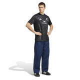 A person stands with hands on hips, wearing a black All Blacks Greatest Rivalry Supporters Tee by adidas—a tribute to New Zealand rugby—loose blue pants, and black adidas shoes, against a plain white background.