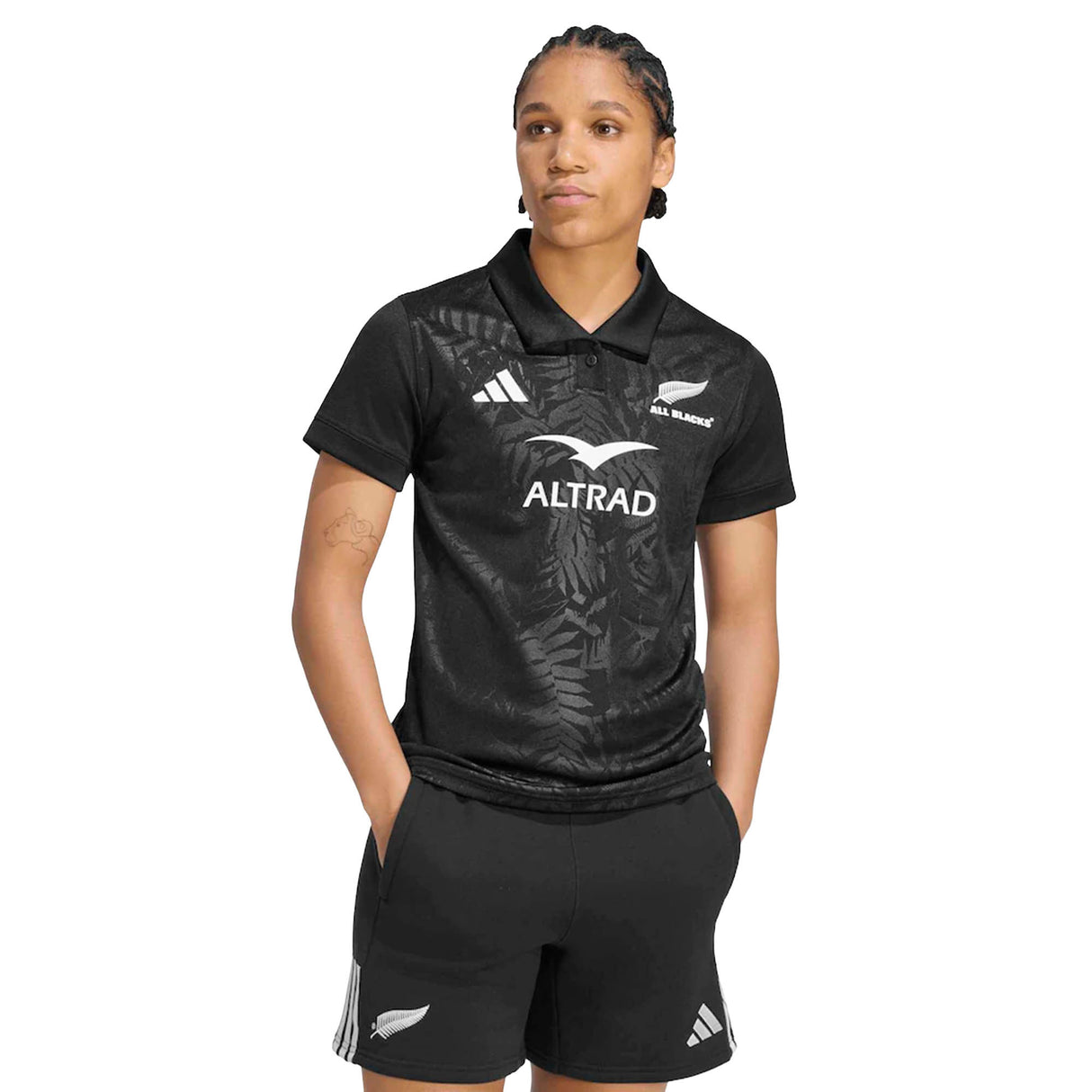 A person wearing the adidas Women’s All Blacks Greatest Rivalry Replica Home Jersey, paired with matching shorts, stands with hands in pockets and looks to the side.