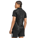 A person with braided hair is seen from behind wearing the adidas Women’s All Blacks Greatest Rivalry Replica Home Jersey—featuring a black collared design with a subtle leaf pattern—and black shorts with white stripes.