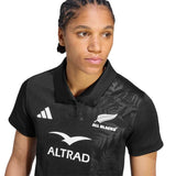 A person wearing the Women’s All Blacks Greatest Rivalry Replica Home Jersey by adidas—a black rugby polo shirt featuring the iconic adidas logo—looking slightly to the side.