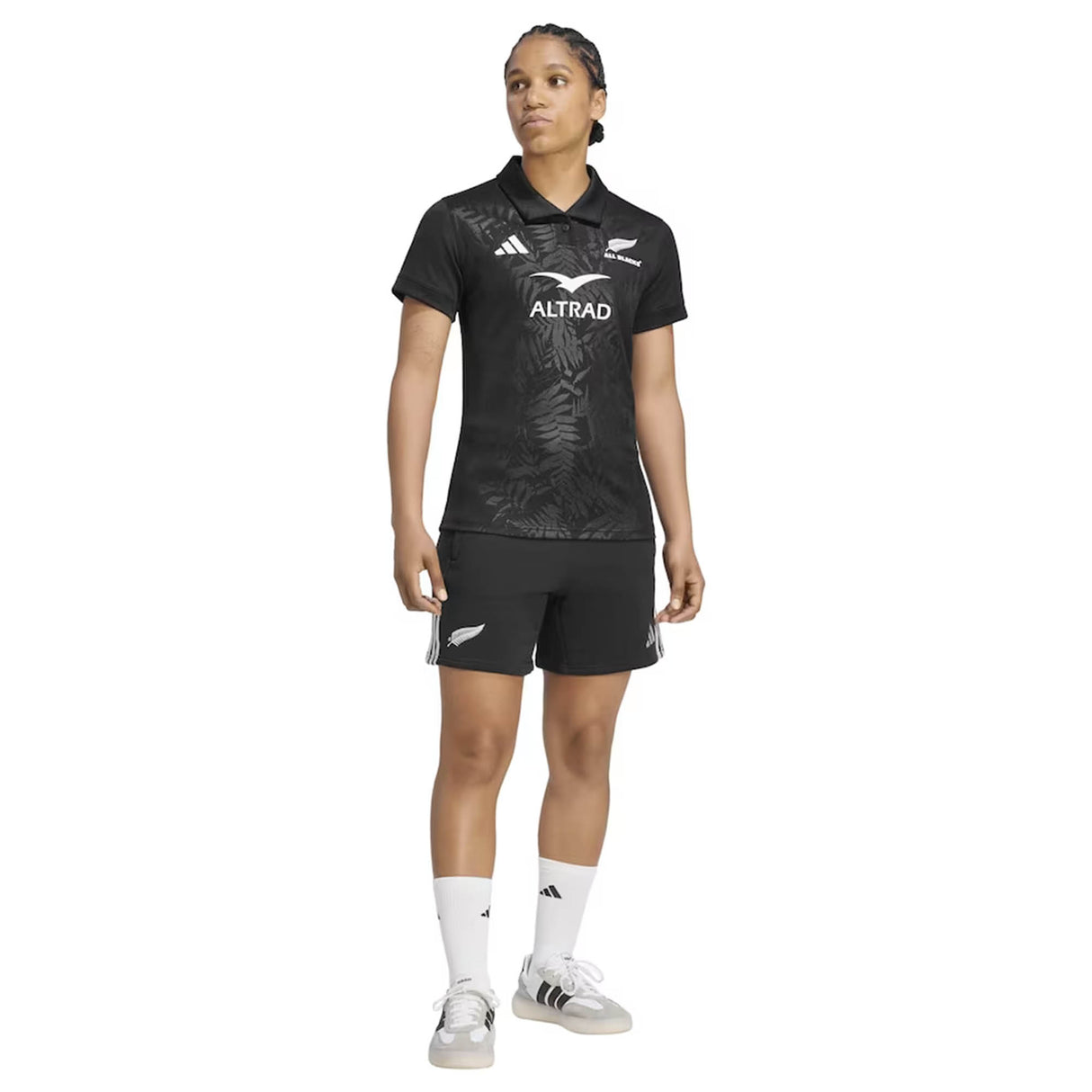 A person is standing in the Women’s All Blacks Greatest Rivalry Replica Home Jersey by adidas and matching shorts, with white socks and shoes. The black jersey and shorts feature adidas sports logos, and the person is looking slightly to the right.