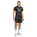 A person is standing in the Women’s All Blacks Greatest Rivalry Replica Home Jersey by adidas and matching shorts, with white socks and shoes. The black jersey and shorts feature adidas sports logos, and the person is looking slightly to the right.