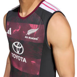 A person wearing the All Blacks Greatest Rivalry Training Singlet by adidas, a sleeveless rugby jersey featuring Toyota and adidas three-stripe logos, with maroon and black patterns inspired by the South Africa tour.