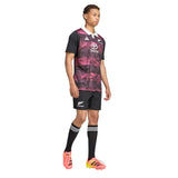 A person stands wearing the adidas All Blacks 26/27 Training Jersey – South Africa Tour, paired with black shorts, black knee-high socks with white stripes, and bright orange cleats against a white background—a look that echoes the official kit for the South Africa tour.