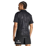 A person with braided hair stands with their back to the camera, wearing the All Blacks Greatest Rivalry Performance Home Jersey by adidas and matching black shorts featuring the Gallagher logo, evoking the iconic style of a classic All Blacks jersey.
