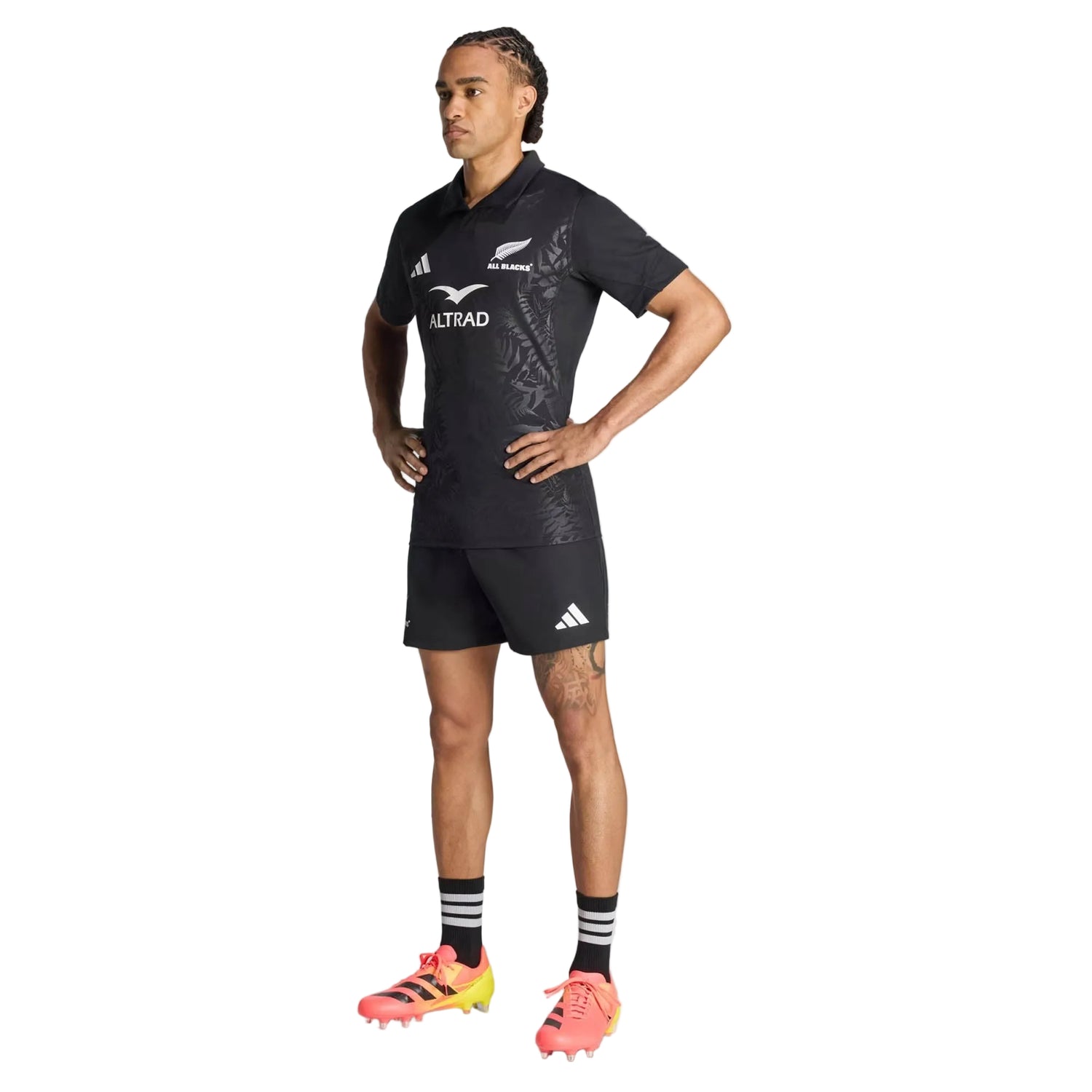 An individual stands with hands on hips, wearing the All Blacks Greatest Rivalry Performance Home Jersey by adidas, paired with black shorts, striped socks, and pink cleats, against a plain white background.