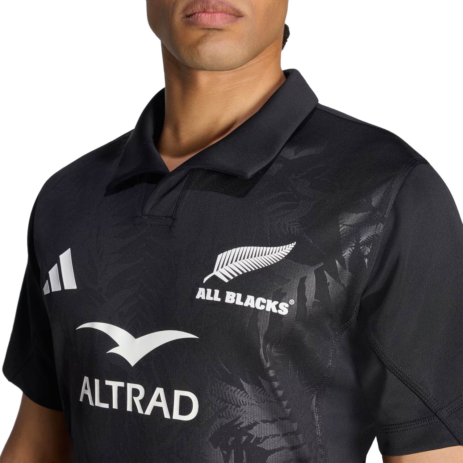 A person wearing the adidas All Blacks Greatest Rivalry Performance Home Jersey, featuring “All Blacks” text, a silver fern logo, and “ALTRAD” sponsor branding; only the upper torso and part of the face are visible.