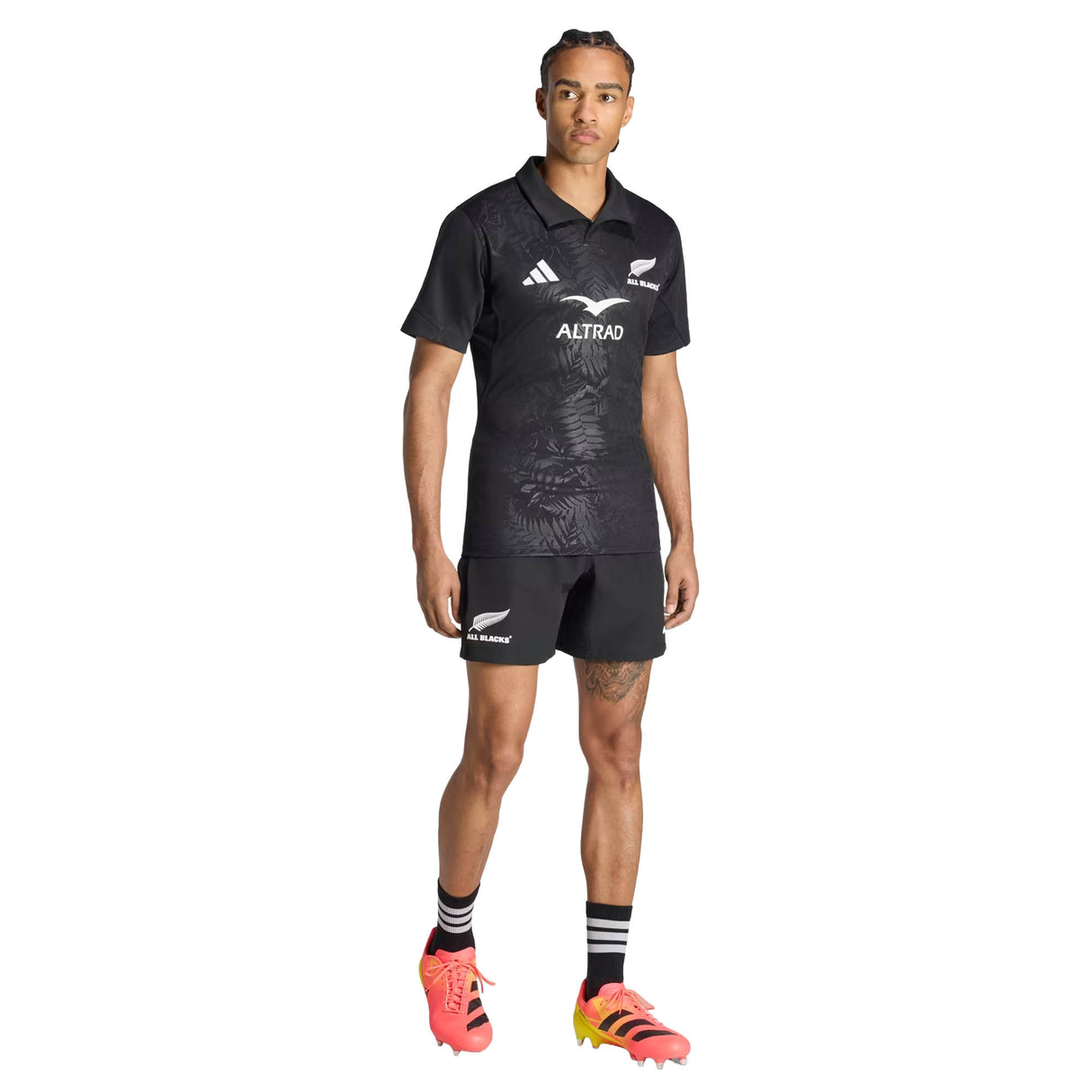 A person stands facing forward against a plain white background, wearing the All Blacks Greatest Rivalry Performance Home Jersey by adidas, along with matching black shorts, striped socks, and orange-yellow cleats.