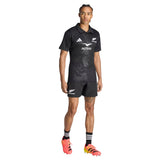 A person stands facing forward against a plain white background, wearing the All Blacks Greatest Rivalry Performance Home Jersey by adidas, along with matching black shorts, striped socks, and orange-yellow cleats.