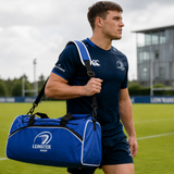 A rugby player in a Leinster Rugby kit walks across a grassy field by a modern building, carrying the Castore Leinster Duffle Bag/Holdall 24/25.