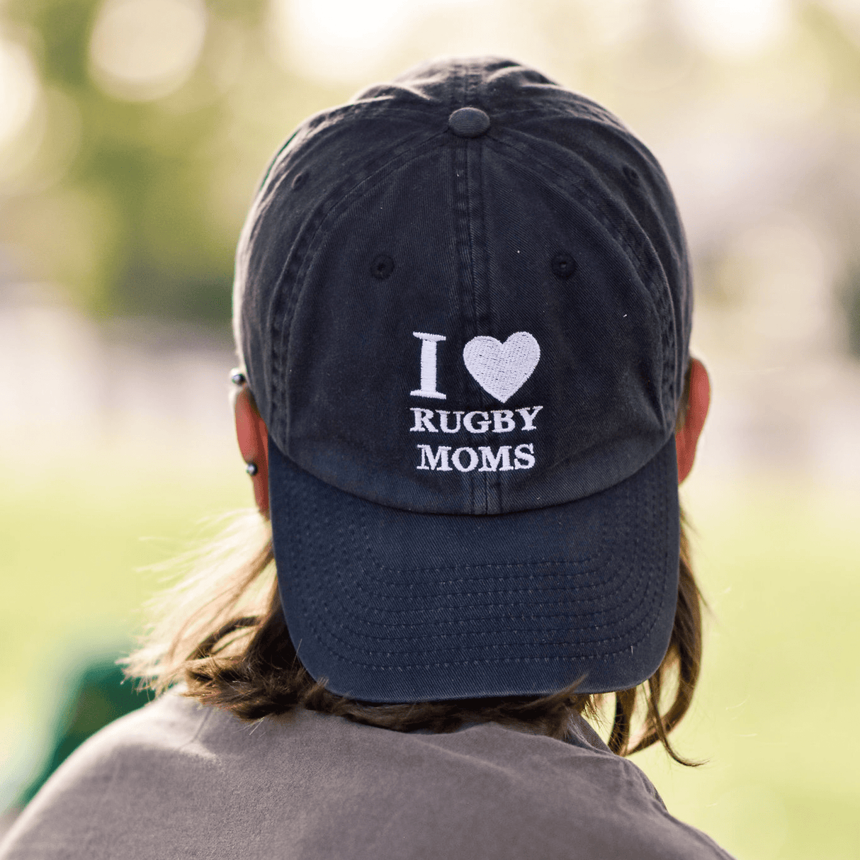 Close-up of the back of a person's head wearing a navy embroidered I Heart Rugby Moms Cap by Modish Rugger, against a blurred green background.
