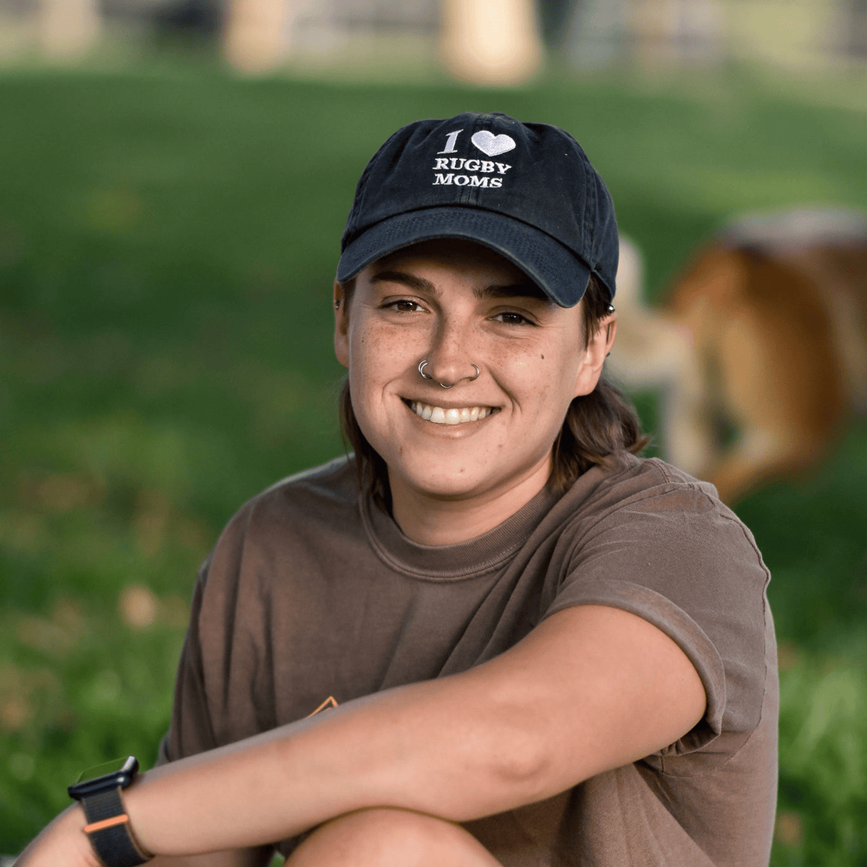 A smiling young woman wearing an "I Heart Rugby Moms Cap" by Modish Rugger, sitting outdoors with a blurred green background and a partial view of a dog.