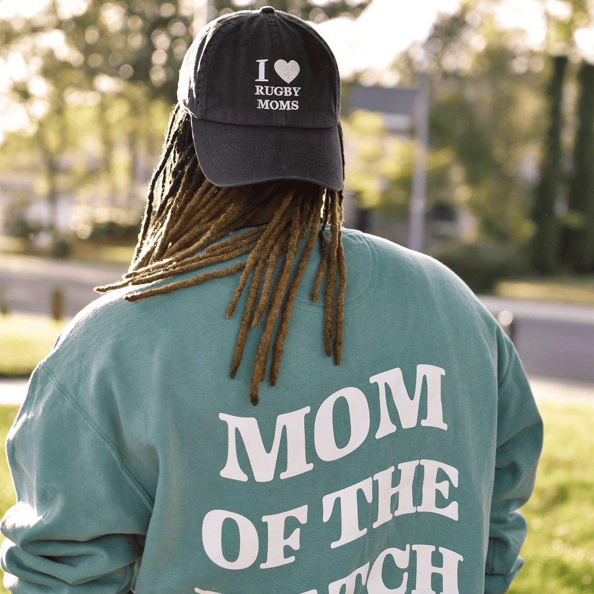 Person with dreadlocks wearing a teal sweatshirt that says "mom of the match" and an I Heart Rugby Moms Cap by Modish Rugger, viewed from the back.