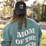Person with dreadlocks wearing a teal sweatshirt that says "mom of the match" and an I Heart Rugby Moms Cap by Modish Rugger, viewed from the back.