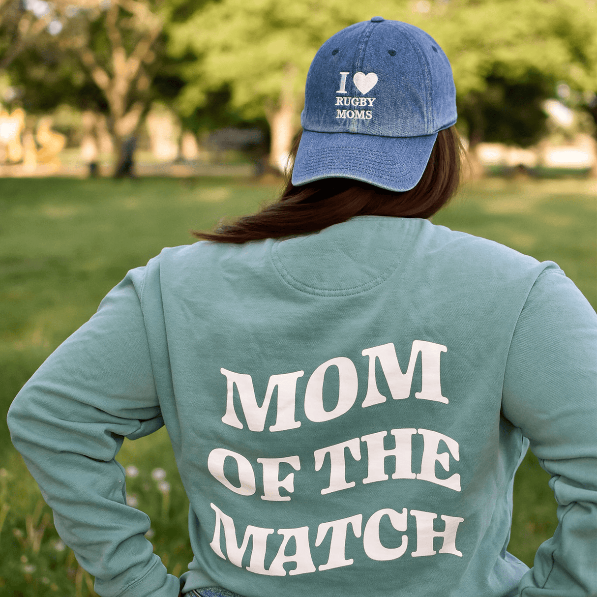 Woman in a park wearing a green sweatshirt reading "Rugby Mom Pride" and an I Heart Rugby Moms Cap by Modish Rugger.