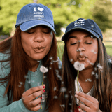 Two women wearing "I Heart Rugby Moms" caps by Modish Rugger blow dandelions, with seeds dispersing in the air, in a sunlit park.